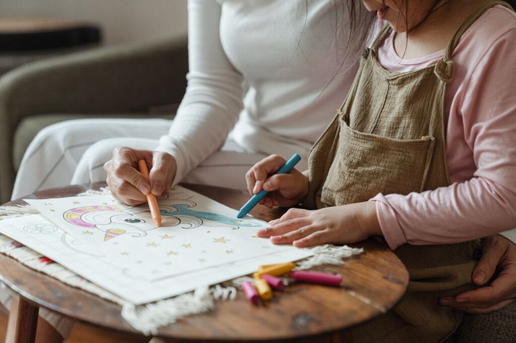 Side view of crop anonymous woman and little girl in casual clothes sitting on sofa and coloring drawing with wax colored pencils on coffee table
