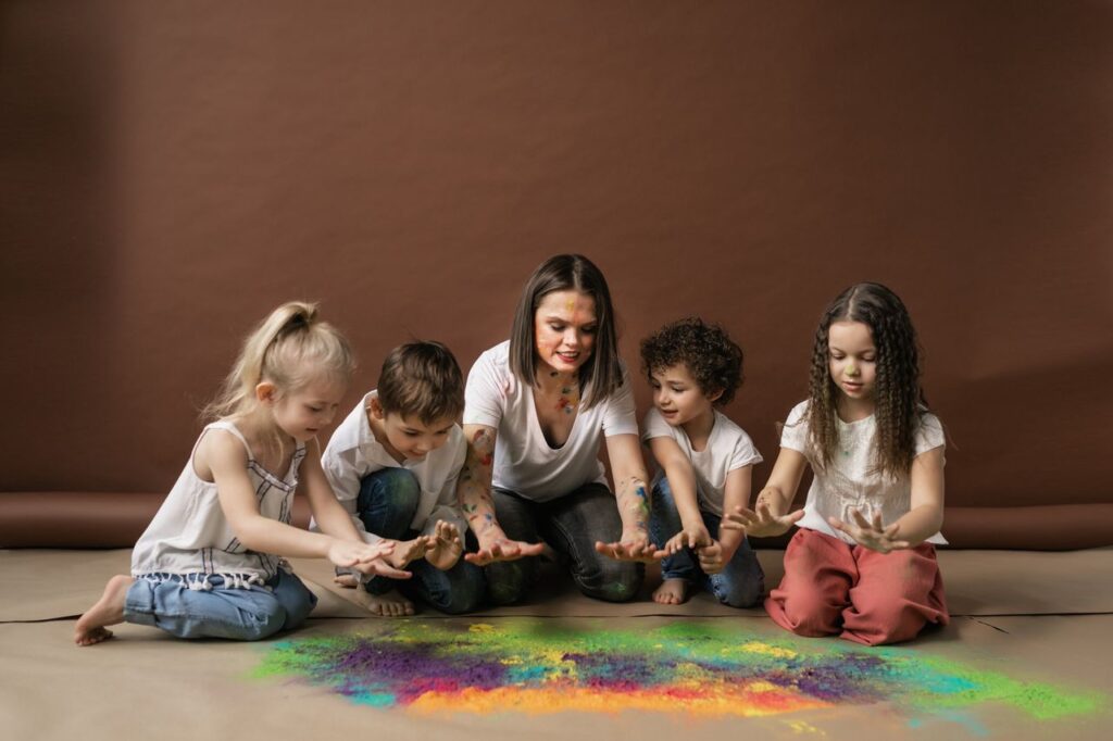 Group of children and a woman enjoying colorful powders together indoors, expressing fun and creativity.